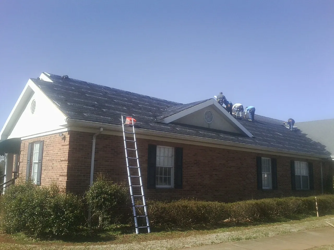 Crew tearing shingles off a brick ranch roof with a ladder set — Metal Roof Repair work by our crew in Browns Summit